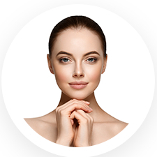 A person with fair skin and dark hair is looking directly at the viewer, smiling slightly, with hands gently touching their chin. The background is white, and the image is circular.